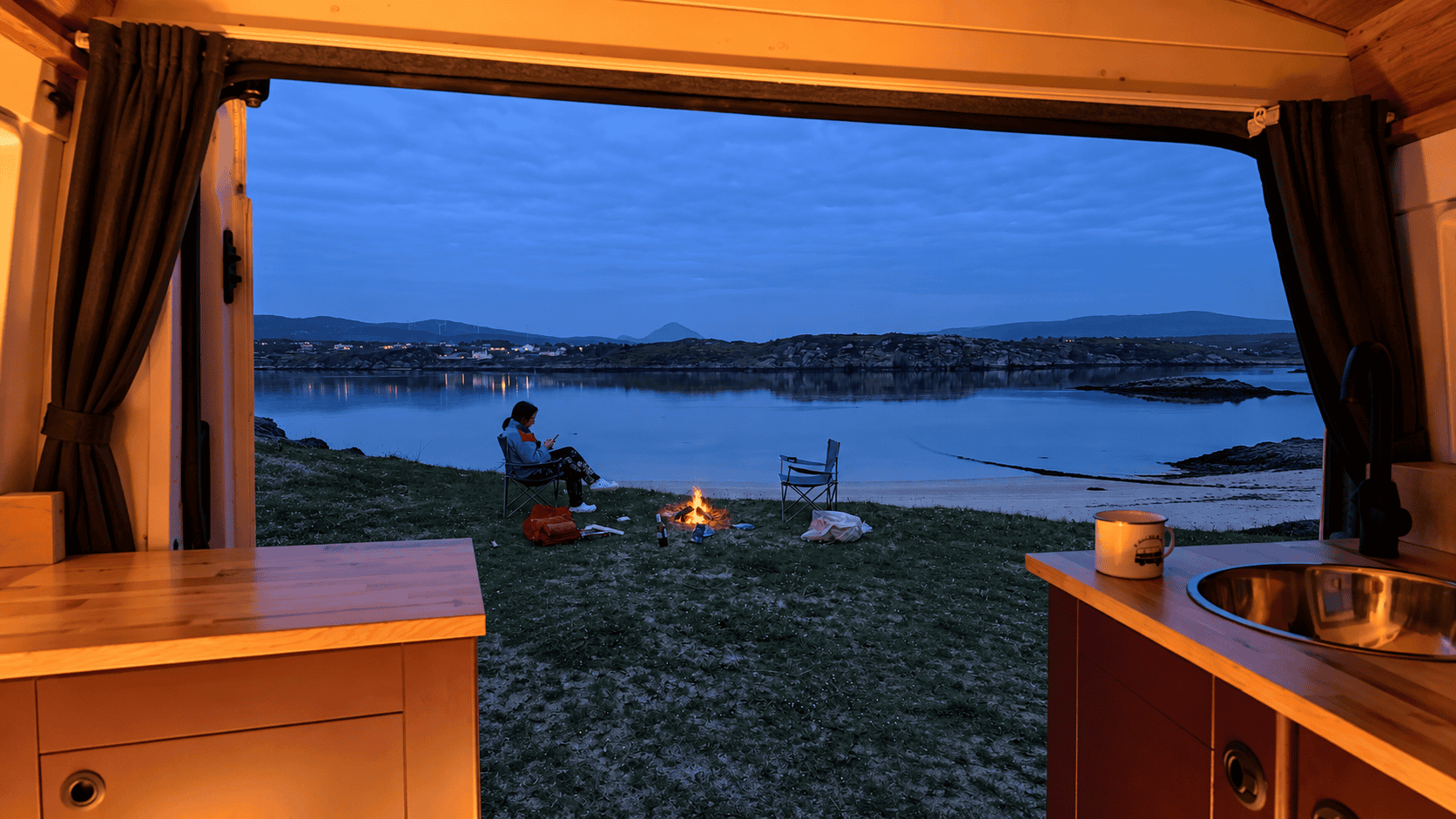 Warm wooden campervan interior looking out towards a person beside a small fire at dusk.